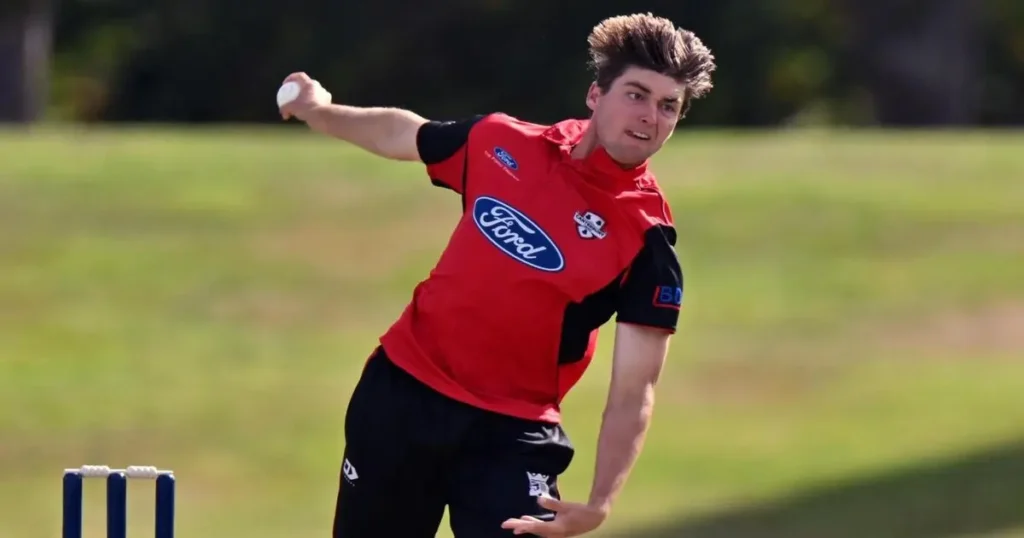 New Zealand pacer Zakary Foulkes mid-action during a delivery stride, wearing a red and black Canterbury kit against a green field backdrop.